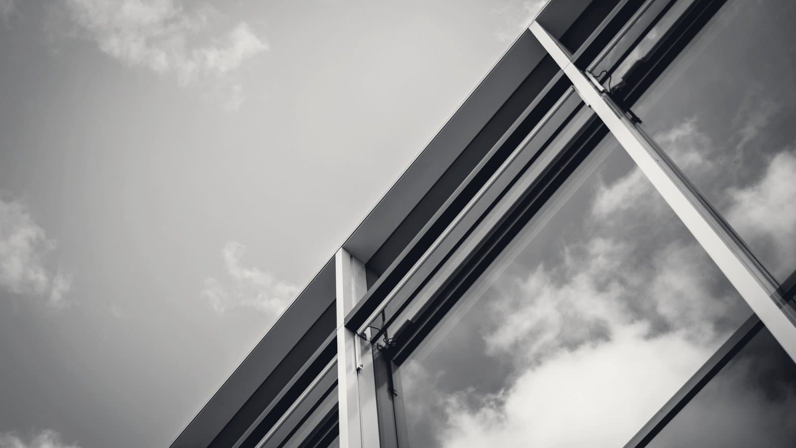A glass curtain wall angled upward against an overcast sky, in monochrome.