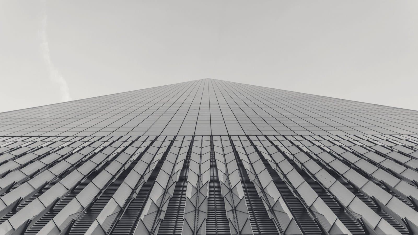 A glass tower photographed from its base, its facade tapering to a single point against a pale sky.